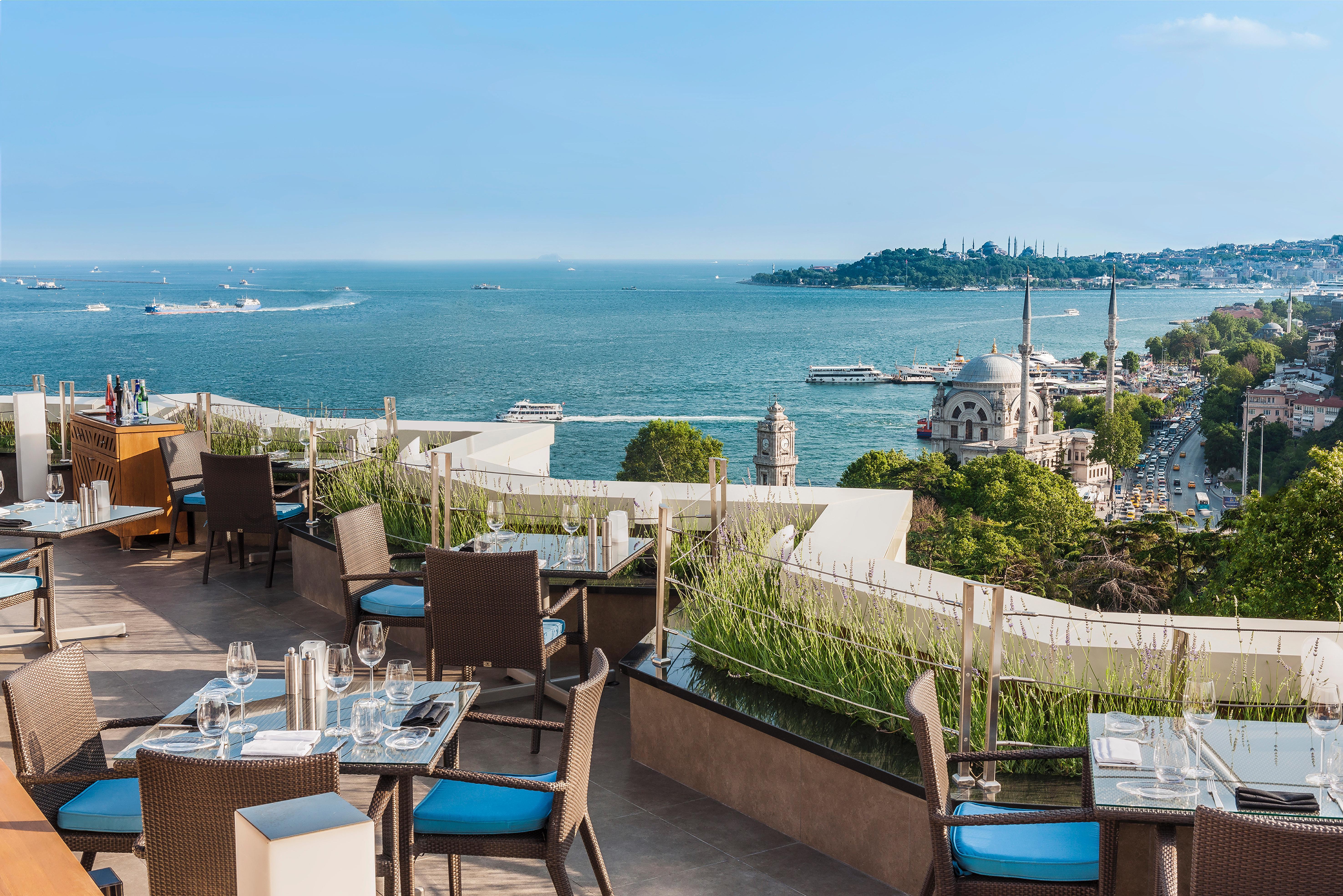 Swissotel The Bosphorus Istanboel Buitenkant foto The image shows a beautiful outdoor dining area on a terrace overlooking a scenic coastal view. The setup includes several tables with elegant dining arrangements, featuring chairs with blue cushions. In the background, there is a vast body of water,