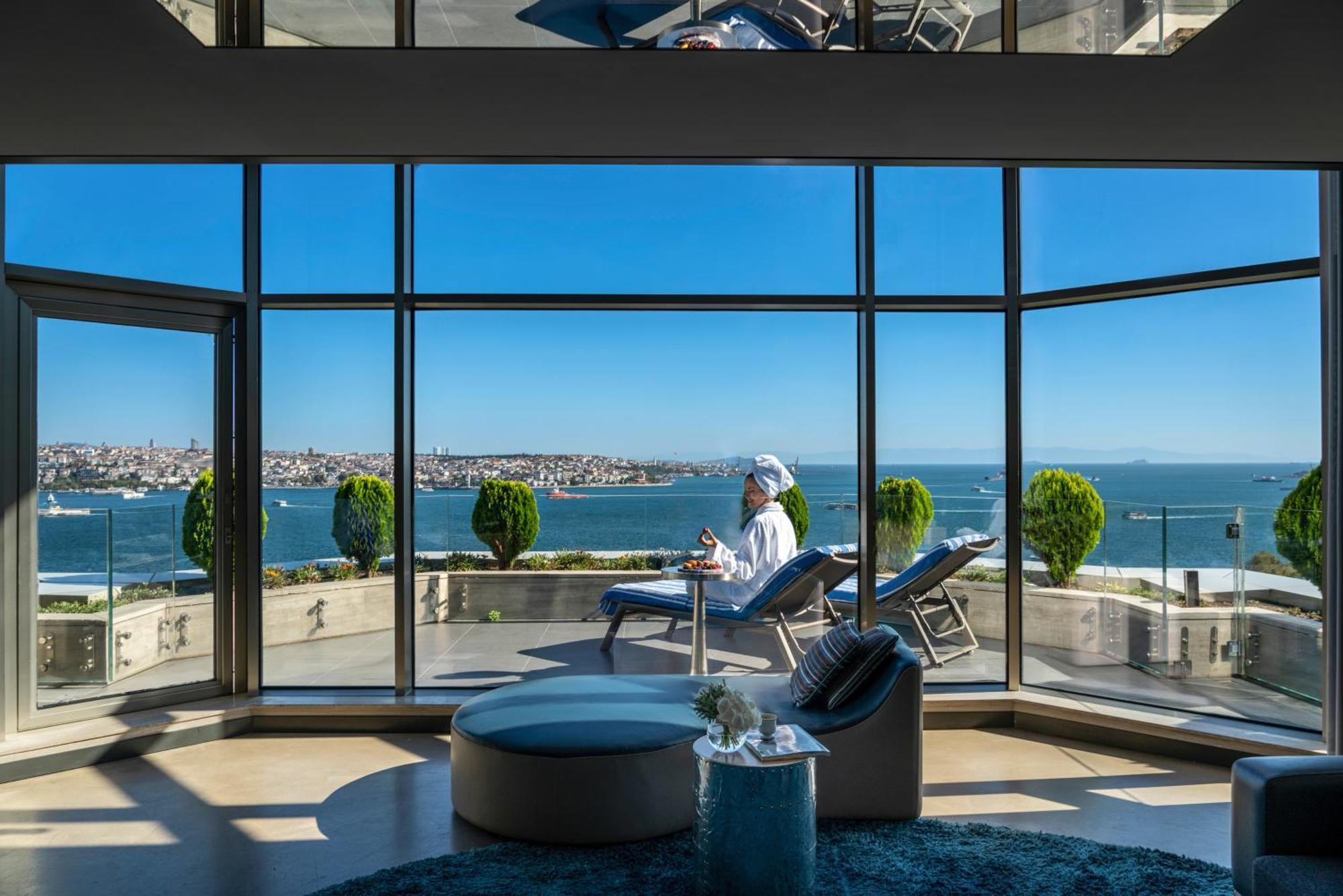 Swissotel The Bosphorus Istanboel Buitenkant foto The photo shows a luxurious indoor setting with large glass windows providing a panoramic view of a body of water and distant cityscape. Inside, there is a person sitting comfortably, likely in a leisure pose, wearing a robe or relaxed clothing. The 