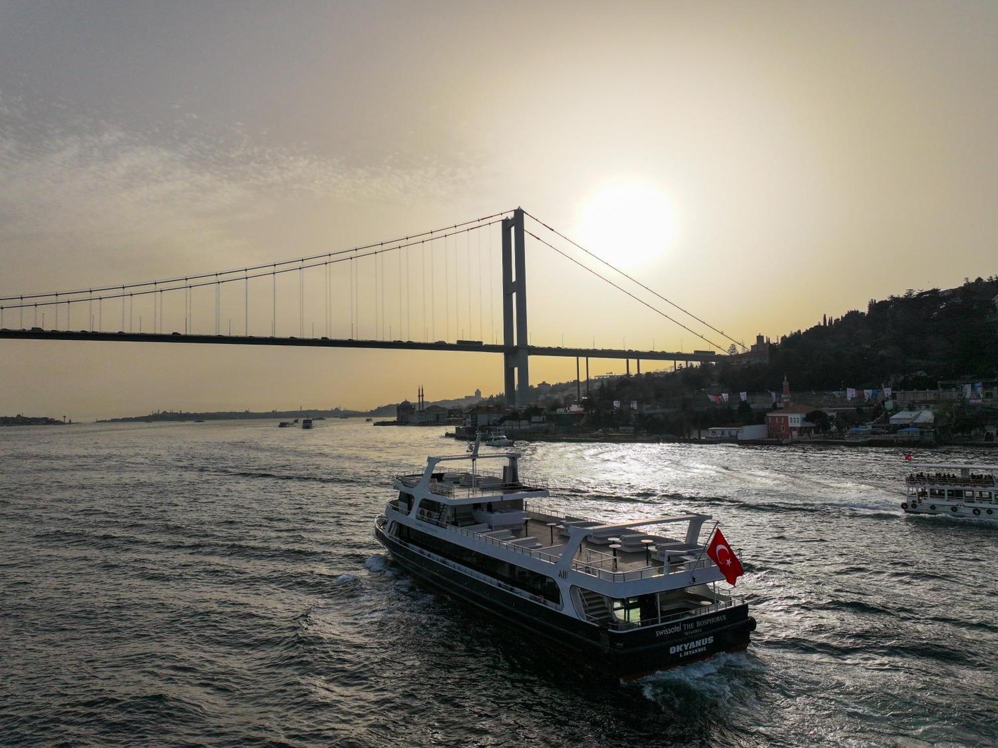 Swissotel The Bosphorus Istanboel Buitenkant foto The photo depicts a scenic view of a large body of water with a ferry boat traveling across it. The ferry is adorned with a Turkish flag. In the background, there is a bridge spanning the water, with the sun setting or rising behind it, casting a war