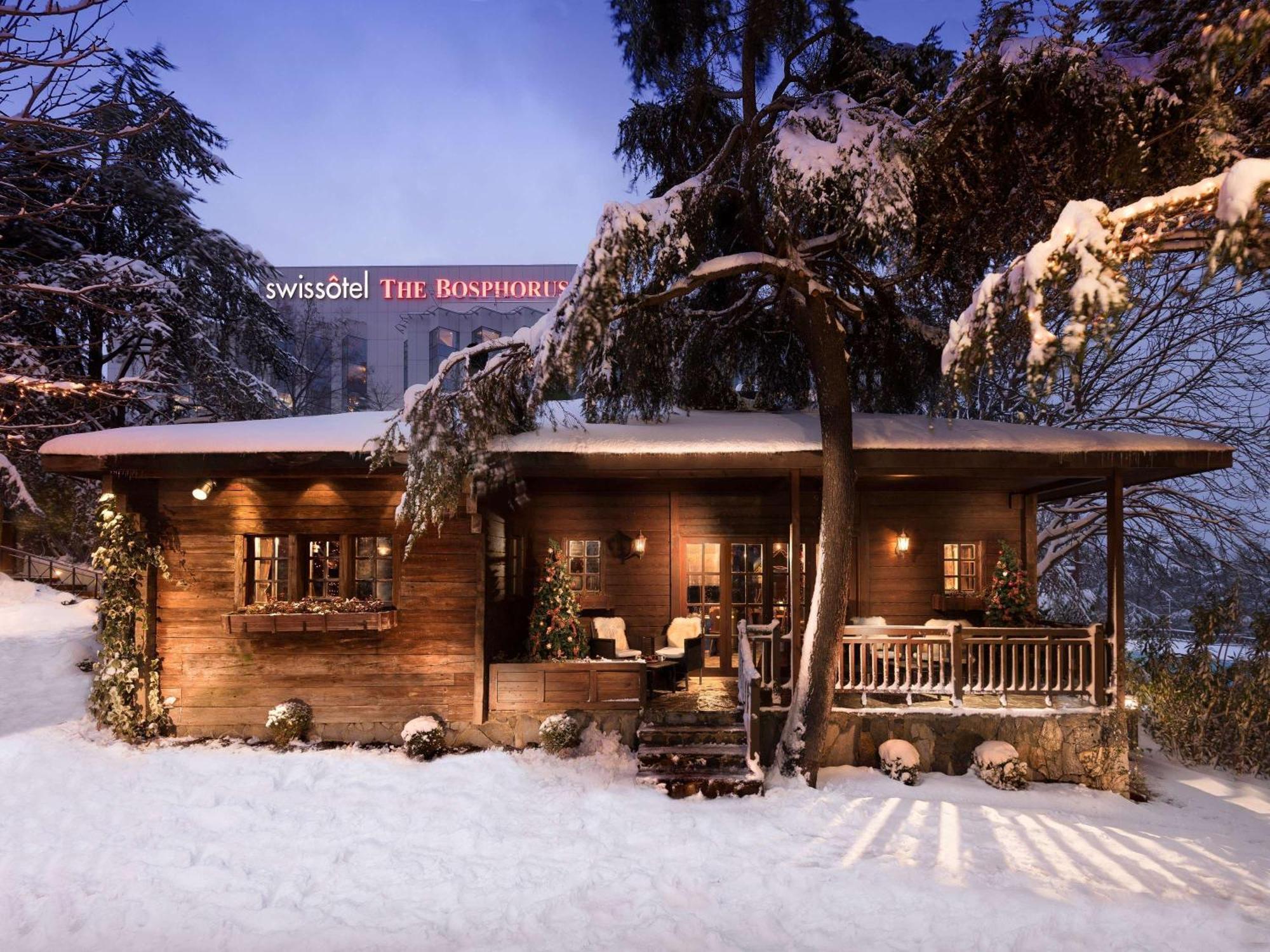 Swissotel The Bosphorus Istanboel Buitenkant foto The photo shows a charming wooden cabin or chalet nestled in a snowy landscape. The structure features a cozy design with a deck and seating area out front, surrounded by trees. In the background, there is a prominent building with the name "Swissôte